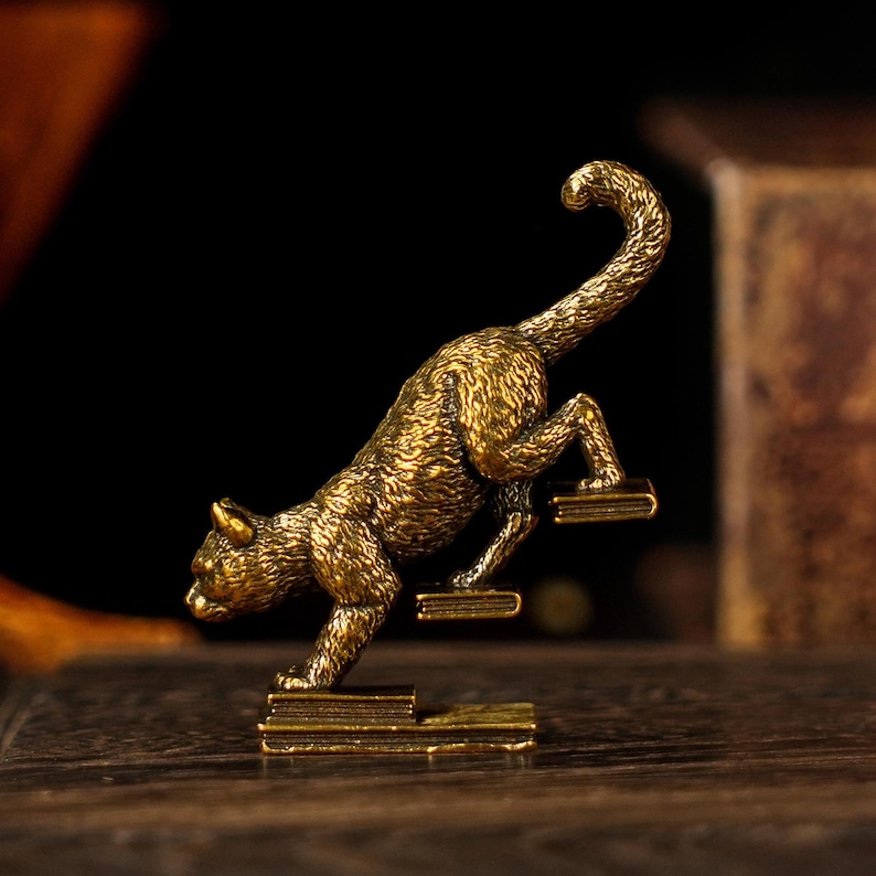 May include: A detailed, golden-colored cat figurine climbing a set of miniature steps. The cat is in a dynamic pose, with its tail curved upwards. The figurine is made of metal and has a textured surface, suggesting a bronze or brass material.