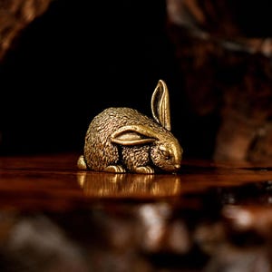 Op de afbeelding: Een kleine, gedetailleerde messing konijnenfiguur. Het konijn zit, met één oor omhoog. Het oppervlak van het konijn is getextureerd en de algehele kleur is warm goud. Het beeldje staat op een houten oppervlak.