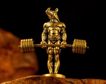 May include: A gold-colored figurine of a muscular rhino lifting a barbell. The rhino is posed in a weightlifting stance, with the barbell held at chest level. The figurine is detailed and stands on a wooden base.