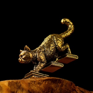 May include: A bronze cat figurine climbing a set of two book-shaped steps. The cat is detailed with textured fur and a raised tail. The figurine is set against a dark background, highlighting the golden color of the sculpture.