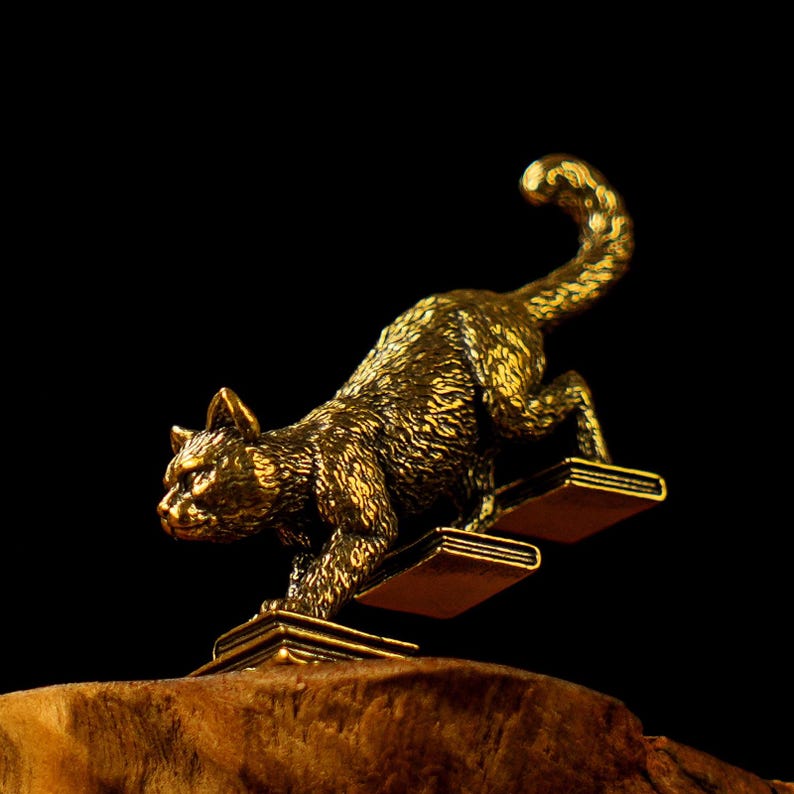 May include: A bronze cat figurine climbing a set of two book-shaped steps. The cat is detailed with textured fur and a raised tail. The figurine is set against a dark background, highlighting the golden color of the sculpture.