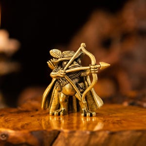 Könnte beinhalten: Eine kleine, goldfarbene Messingfigur eines Frosches, der als Bogenschütze gekleidet ist. Der Frosch trägt einen Hut mit Feder, einen Umhang und einen Gürtel mit Taschen und hält Pfeil und Bogen mit beiden Händen in Schussposition.