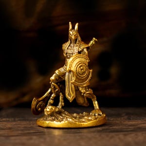 May include: A small, gold-coloured Anubis figurine. The Egyptian god is depicted in a dynamic pose, holding a shield and a staff. The figure has a detailed design and is mounted on a circular base. The background is a blurred, dark brown.