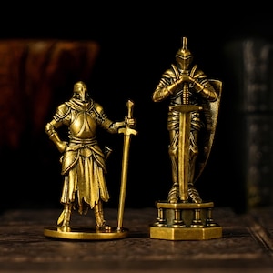 May include: Two brass knight figurines. One knight stands with a sword at his side, the other holds a sword and shield. Both are clad in detailed armor, with helmets and decorative bases. The figurines are set against a dark background.