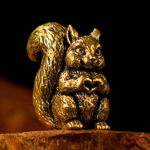 May include: A detailed golden squirrel figurine with a heart-shaped gesture. The squirrel is sitting upright, with its paws forming a heart. The figurine has a textured surface and is set against a dark background.