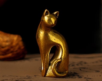 May include: A small, golden cat figurine in a seated pose. The cat has a curved back, pointed ears, and a curled tail. The figurine is made of a shiny, metallic material and sits on a patterned surface. The background is dark.
