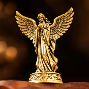 May include: A metallic gold-toned angel figurine standing on a circular base with decorative engravings. The angel has large, detailed wings spread wide, long flowing robes, and hands clasped together near the chest in a prayer-like position.