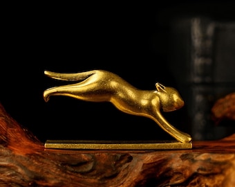 May include: A gold-colored metal sculpture of a leaping cat. The cat is in mid-air, with its body arched and legs extended. The sculpture is mounted on a rectangular base and set against a dark background.