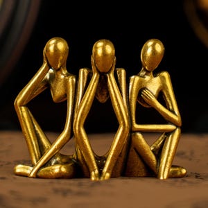 以下が含まれることがあります： 金色の彫刻で、目、耳、口をそれぞれ手で覆っている3人の座っている人物を描いています。人物は暗い背景の茶色の表面にいます。彫刻は装飾品です。