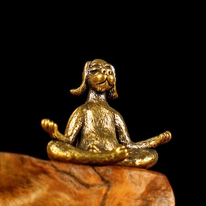 Puede incluir: Una figurita de metal de color dorado de un perro sentado en una postura de yoga. El perro tiene los ojos cerrados y la boca ligeramente abierta. La figurita está sentada sobre un trozo de madera.