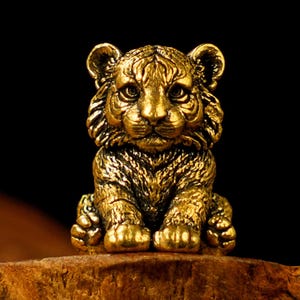 Può includere: Una dettagliata statuetta di cucciolo di tigre color oro. La scultura presenta intricati dettagli della pelliccia, dei lineamenti del viso e delle zampe della tigre. Il cucciolo è seduto in posizione eretta, con le zampe anteriori appoggiate su una superficie di legno, su sfondo nero.