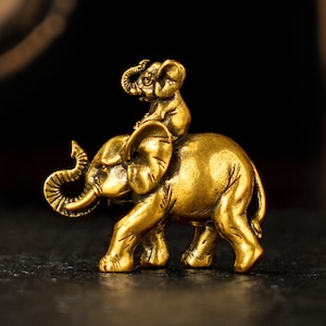 May include: A small, detailed brass figurine of an elephant with a smaller elephant riding on its back. The figurine has a polished, golden finish and is set against a dark background. The elephant's trunk is curled upwards, and the smaller elephant is perched on its head.