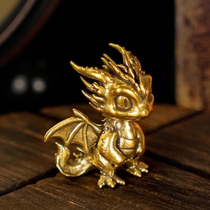 May include: A small, detailed brass dragon figurine with wings, horns, and a curled tail. The dragon is posed in a seated position, with intricate detailing on its scales and features. The figurine is displayed on a wooden surface.
