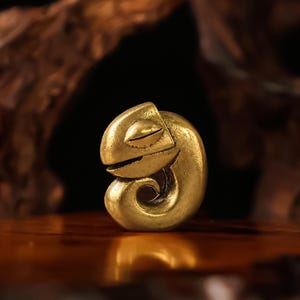 May include: A golden, stylized chameleon figurine in a curled pose. The sculpture has a smooth, polished finish and closed eyes, suggesting a relaxed or sleeping state. The chameleon's tail curls into a spiral, adding a decorative element.