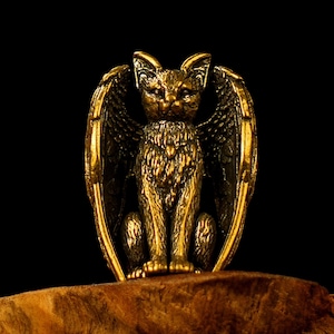 May include: A golden cat figurine with large, feathered wings, set against a black backdrop. The cat is depicted in a seated position, with detailed fur and wing textures. The statue's metallic finish is prominent, showcasing its intricate design.