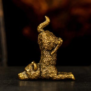 May include: A small, detailed brass figurine of a bear in a yoga pose. The bear is on its knees with its back arched and tail curled. The figurine has a textured surface and is a warm gold colour.