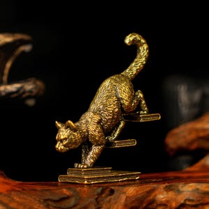 May include: A detailed brass cat figurine in a climbing pose. The cat is depicted with textured fur and is positioned on a stack of books. The sculpture has a golden hue and is set against a dark background.