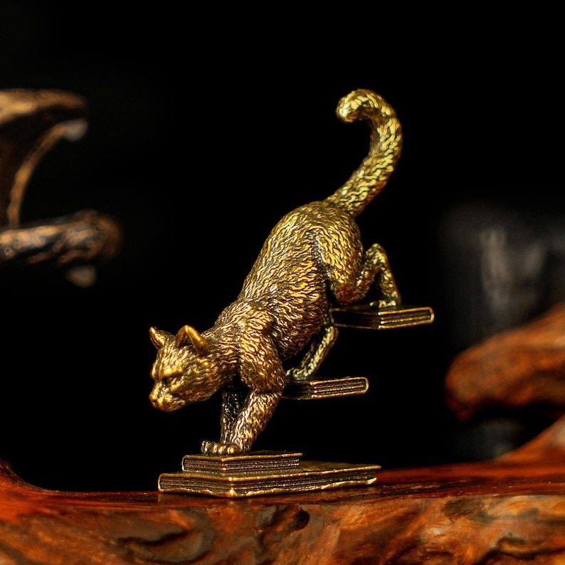 May include: A detailed brass cat figurine in a climbing pose. The cat is depicted with textured fur and is positioned on a stack of books. The sculpture has a golden hue and is set against a dark background.