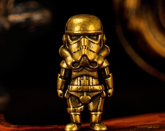 May include: A small, detailed brass figurine of a Stormtrooper from Star Wars. The figure is in a standing pose, with a helmet and armor. The brass has a polished, golden finish. The background is dark.