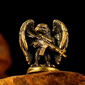 Peut inclure: Figurine détaillée de couleur bronze représentant un aigle tenant un fusil. L'aigle a les ailes déployées et se tient sur une petite base ronde. La figurine est très détaillée, avec des détails complexes de plumes et d'armes.
