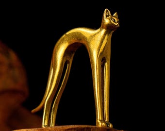 May include: A golden cat figurine with an arched back and slender legs. The cat is posed in a standing position, with its head turned slightly to the side. The figurine is made of a shiny, metallic material and is set against a dark background.
