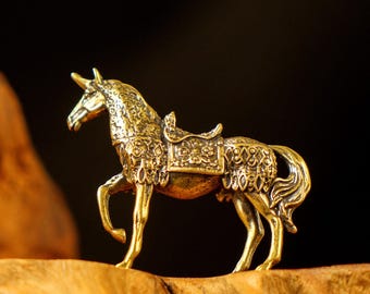 May include: A detailed, golden-colored figurine of a horse with a saddle. The horse is posed mid-stride, with intricate patterns adorning its body and saddle. The figurine is set against a dark background, highlighting its metallic sheen.
