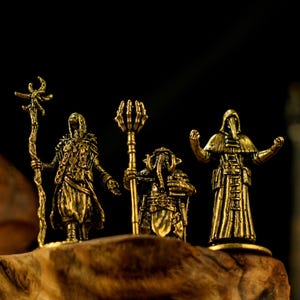 Könnte beinhalten: Drei antike, goldfarbene Figuren von Fantasy-Charakteren. Die Figuren umfassen eine Figur mit Robe und Stab, ein Wesen mit Stab und eine Pestdoktorfigur. Die Figuren werden auf einer Holzoberfläche ausgestellt.