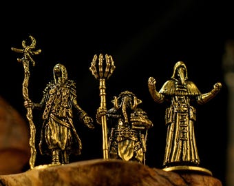 Puede incluir: Tres figuras antiguas de color dorado de personajes de fantasía. Las figuras incluyen un personaje con túnica y báculo, una criatura con báculo y una figura de médico de la peste. Las figuras se exhiben sobre una superficie de madera.