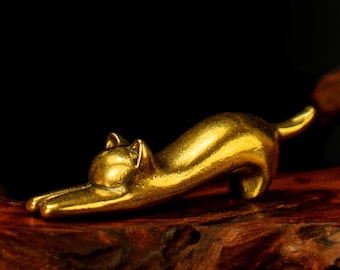 Può includere: Piccola statuetta dorata a forma di gatto in posizione di allungamento. Il gatto è realizzato in un materiale metallico lucido e si trova su una superficie di legno. Lo sfondo è scuro, evidenziando la forma del gatto.