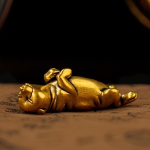 May include: A small, gold-coloured figurine of a reclining animal, possibly a dog or a pug, with its paws raised. The figurine has a smooth, polished surface and is lying on its back. The background is dark.