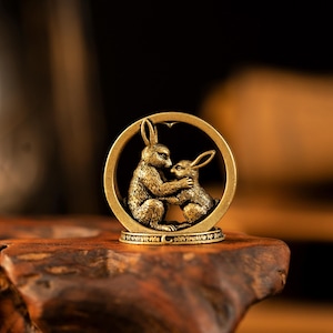 May include: A brass-coloured metal ornament featuring two rabbits facing each other within a circular frame. The rabbits are depicted in a seated position with textured fur details. The circular frame rests on a small, flat, decorative base.