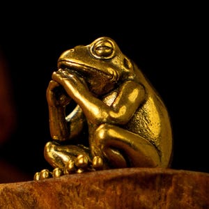 Puede incluir: Una figura de rana dorada en posición sentada, con las manos juntas cerca de la cara. La rana tiene una expresión contemplativa, con los ojos parcialmente cerrados. La figura está sobre un fondo oscuro, resaltando su brillo metálico.