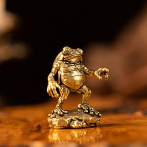 May include: A small, metallic gold-colored frog figurine standing in an upright, anthropomorphic pose. The frog has textured skin, prominent eyes, and extended limbs, and it is mounted on a small, uneven base that mimics a rocky surface.