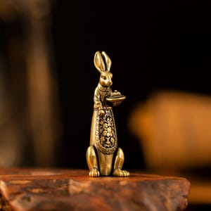 May include: A brass figurine of an upright rabbit wearing a collared shirt and an apron with a detailed floral pattern. The rabbit is holding a small, round tray in its front paws. The metal has a warm, golden-brown finish with dark patina.