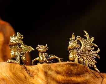 Puede incluir: Tres figuras de dragones de latón en varias poses se encuentran sobre un trozo de madera. Los dragones tienen alas, escamas y cuernos detallados. La madera tiene un aspecto natural y texturizado, y el fondo es oscuro.