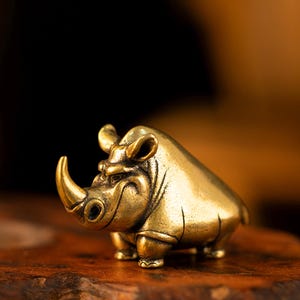 May include: A small, stylized brass rhinoceros figurine with a polished, metallic gold finish. The sculpture features a prominent horn, rounded ears, and a stout body with visible etched lines defining the legs and facial features of the animal.