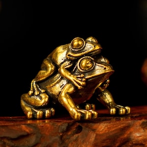 Puede incluir: Pequeña y detallada figura de rana de latón. Una rana está posada sobre la espalda de otra, ambas mirando hacia adelante. Las ranas tienen ojos grandes y piel texturizada, y el diseño general es de color oro cálido.