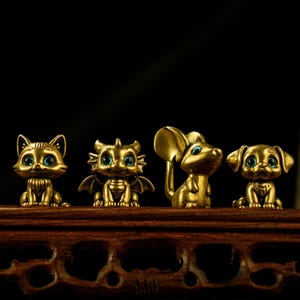 May include: Four small, gold-colored animal figurines: a cat, a dragon, a mouse, and a dog. Each figure has large, blue eyes. They are displayed on a dark wooden surface against a black background.