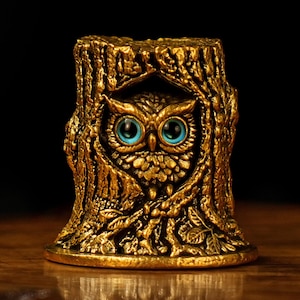 Könnte beinhalten: Goldfarbene Eulenfigur mit leuchtend blauen Augen, eingebettet in einen detaillierten Baumstumpf. Die Eule ist aufwendig gestaltet, mit strukturierten Federn und einem fokussierten Blick. Der Sockel ist rund, und der Gesamteffekt ist dekorativ.