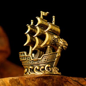 May include: A detailed, gold-coloured pirate ship figurine with three masts and billowing sails. The ship features a skull and crossbones emblem and ornate detailing. The figurine is set against a dark background, highlighting its intricate design.