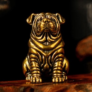 May include: A detailed, golden-colored Shar Pei dog figurine in a seated position. The sculpture has intricate details, including wrinkles and folds, and is set against a dark background. The dog's expression is alert and attentive.