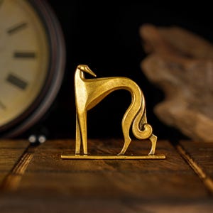 May include: A gold-coloured metal sculpture of a stylised greyhound dog. The sculpture is standing on a small base and has a curled tail.
