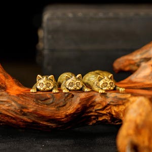 Op de afbeelding: Drie kleine, gouden kattenbeeldjes op een rij, liggend met hun poten uitgestrekt. De katten hebben gedetailleerde kenmerken, waaronder ronde ogen en getextureerde vacht. Ze worden getoond op een stuk gepolijst, donkerbruin hout.