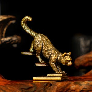 May include: A detailed brass cat figurine climbing a stack of books. The cat is depicted in a dynamic pose, with its tail curved and body angled. The figurine has a textured surface and is set against a dark background.