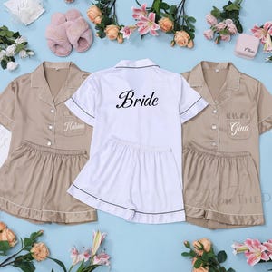 May include: Tan satin pajama sets with white piping and personalized names, alongside a white set with "Bride" in black script. The image includes pink slippers, flowers, and decorative elements, suggesting a bridal theme.