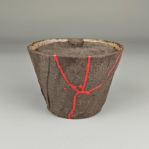 May include: A dark brown ceramic bowl with a lid. The bowl has a textured surface and is decorated with bright red lines. The lid has a small knob on top. The bowl is likely handmade and has a rustic appearance.