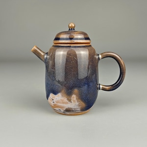 Puede incluir: Una tetera de cerámica con tapa y asa. La tetera tiene un esmalte marrón y azul con efecto craquelado. El asa es un lazo redondeado y el pico es corto e inclinado. La tapa tiene un pequeño pomo en la parte superior.