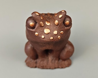 May include: A small, brown ceramic frog figurine with gold accents. The frog has a wide, toothy grin and large, round eyes.