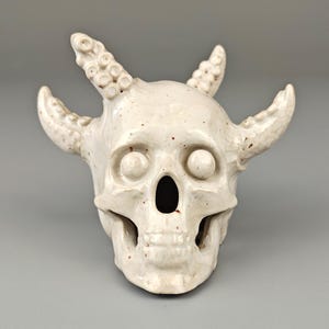 Op de afbeelding: Witte keramische schedel met gedetailleerde, getextureerde hoorns en grote oogkassen. De schedel heeft een gevlekte, verweerde uitstraling.