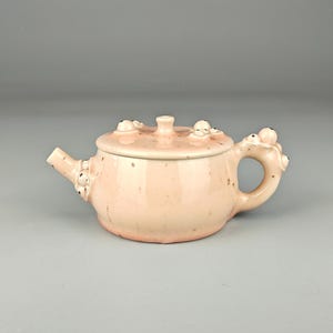 Puede incluir: Pequeña tetera de cerámica rosa con tapa y asa. La tetera tiene un cuerpo redondeado, un pico y un asa de bucle. La tapa está adornada con pequeños elementos decorativos en relieve. La tetera es de color rosa suave con motas más oscuras.